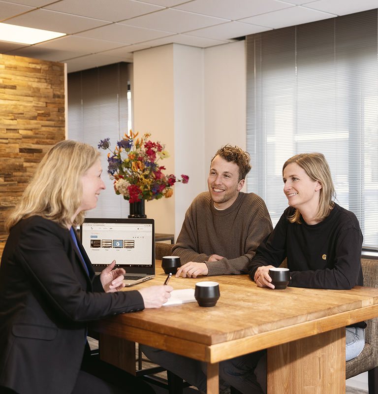 Drie mensen in gesprek aan een houten tafel met een laptop en een vaas bloemen